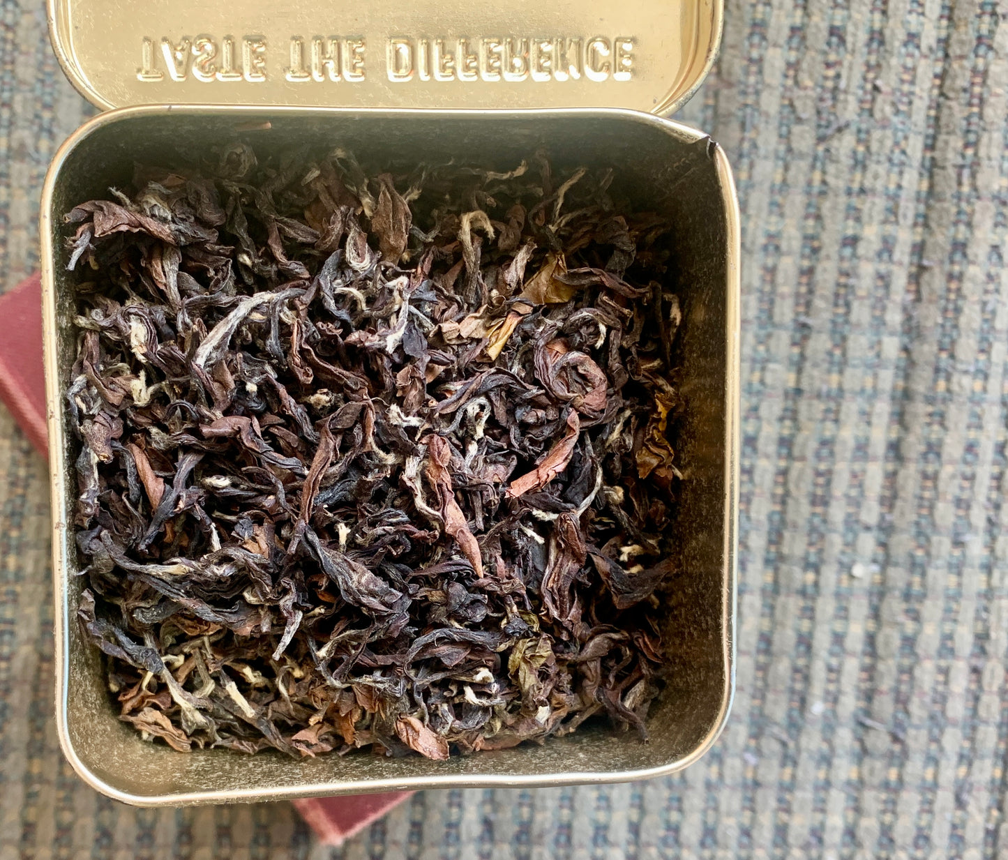 Open tin box with dried tea leaves inside, on a textured surface.