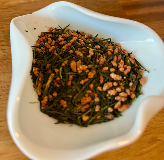 A close-up of loose-leaf green tea mixed with small brown roasted rice, served in a white ceramic dish on a wooden table.