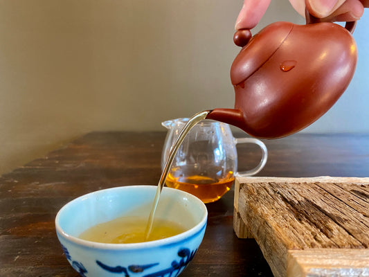 A person pouring tea into a cup with a teapot and a bowl of bubble tea in the foreground, on a table with a wooden plank and a blue vase.