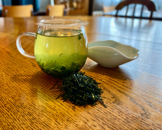 A cup of kabusecha green tea on a wooden table with a pile of green tea leaves in front of it, indicating the tea is freshly brewed and ready to serve.