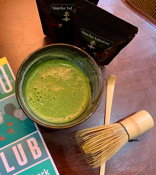 Matcha green tea in a bowl with a whisk and Matcha packaging on a wooden surface.